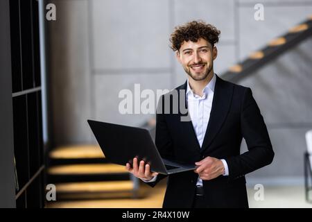 Giovane uomo d'affari moderno che indossa un blazer blu e una camicia casual, in piedi con un laptop aperto in mano Foto Stock