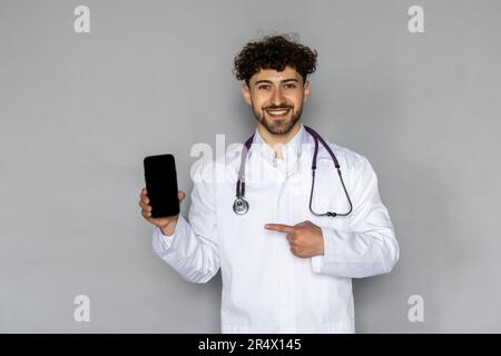 Uomo medico uomo indossa abito bianco medico stetoscopio lavoro in ospedale uso mostrare cellulare schermo vuoto area di lavoro isolato su semplice gr Foto Stock