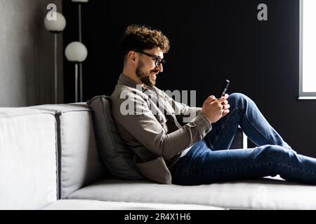 Un uomo serio e premuroso che tocca il mento, guarda lo schermo dello smartphone, seduto sul divano a casa, un giovane uomo d'affari o uno studente che legge cattive notizie Foto Stock