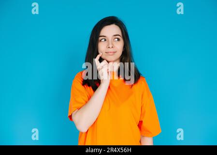 Attraente giovane donna in vestiti arancioni trendy, espressione pensiva e scettica, spazio vuoto per la copia, isolato su sfondo blu - Ritratto foto. Foto Stock