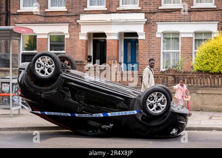 I residenti locali passeggiano davanti alle conseguenze di un incidente d'auto in cui una berlina Nissan capovolta rimane sul lato della strada tra Brixton e Camberwell su Coldharbour Lane, il 30th maggio 2023, a Londra, Inghilterra. Foto Stock