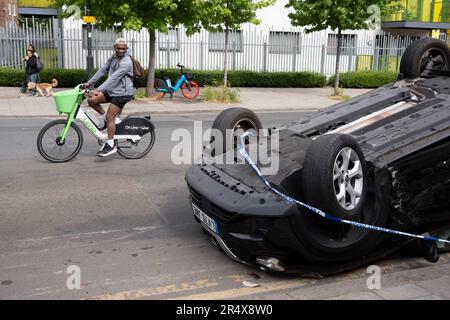 Un ciclista passa le conseguenze di un incidente automobilistico in cui una berlina Nissan ribaltata rimane sul lato della strada tra Brixton e Camberwell su Coldharbour Lane, il 30th maggio 2023, a Londra, Inghilterra. Foto Stock
