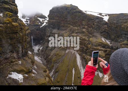 Vista ravvicinata, scattata da dietro, di una donna in piedi e affacciata sul Mulagljufur Canyon, un paradiso per escursionisti, che scatta una foto con il suo smartphone ... Foto Stock