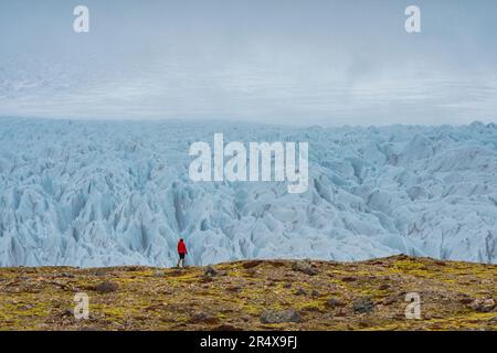 Donna che cammina sulla tundra di fronte a un impressionante ghiacciaio con il suo ghiaccio blu frastagliato e l'atmosfera nebbiosa lungo la costa meridionale dell'Islanda Foto Stock