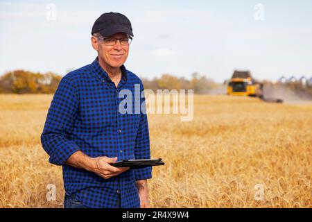 L'agricoltore guarda la telecamera mentre utilizza un tablet per gestire la raccolta della granella con attrezzature di raccolta in background Foto Stock