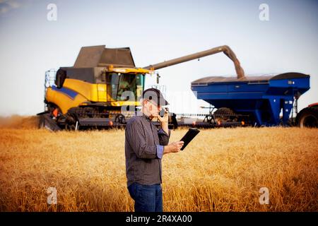 Agricoltore che utilizza un tablet per gestire il raccolto e parlare sul cellulare con una mietitrebbia che si scarica su un carrozzino di grano sullo sfondo Foto Stock