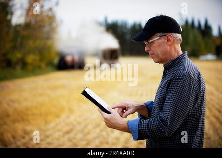 Agricoltore che utilizza un tablet per gestire il raccolto con attrezzature da raccolta in background; Alcomdale, Alberta, Canada Foto Stock