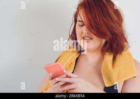 primo piano di una giovane donna latina argentina con capelli rossi corti, con un asciugamano giallo intorno al collo, inviando un messaggio sul telefono su sfondo bianco, Foto Stock