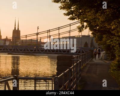 Coppia che cammina lungo la passeggiata sul fiume Odra al tramonto con il ponte di ferro e la cattedrale sullo sfondo; Breslavia, Polonia Foto Stock