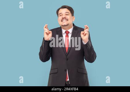 Ritratto di un uomo sorridente soddisfatto con baffi in piedi tiene il dito incrociato, facendo desiderio, pregando per la buona fortuna, indossando vestito nero con cravatta rossa. Studio in interni isolato su sfondo azzurro Foto Stock