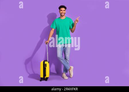 Foto a tutta lunghezza di giovane viaggiatore soddisfatto ragazzo soggiorno con bagaglio diretto dito vuoto spazio nuovo paese isolato su sfondo di colore viola Foto Stock