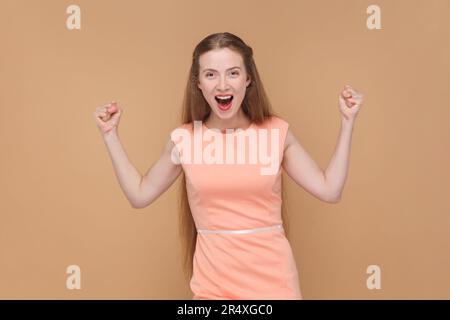 Ritratto di donna estremamente felice positiva con capelli lunghi in piedi con pugni clenched, urlando di eccitazione, indossando un abito elegante. Studio al coperto isolato su sfondo marrone. Foto Stock