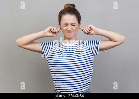 Non voglio sentire questo. Seccato frustrato donna che indossa T-shirt a righe che chiude le orecchie con le dita ignorando le informazioni, stanco di rumore irritante. Studio in interni isolato su sfondo grigio. Foto Stock