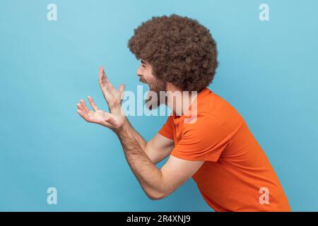 Vista laterale dell'uomo con acconciatura afro che indossa una T-shirt arancione spalancando le mani, guardando seriamente la macchina fotografica, la disputa di famiglia, il malinteso. Studio in interni isolato su sfondo blu. Foto Stock