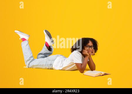 Ragazza curly nera intelligente teen positiva in t-shirt bianca, occhiali legge libro, si trova sul pavimento Foto Stock