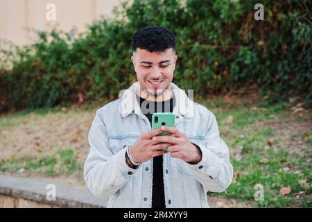Un ragazzo marocchino sorridente che si diverte con uno smartphone per giocare a un videogioco. Messaggi di testo per ragazzi felici sull'applicazione dei social media con un cellulare. Giovane uomo con un telefono cellulare. Foto di alta qualità Foto Stock