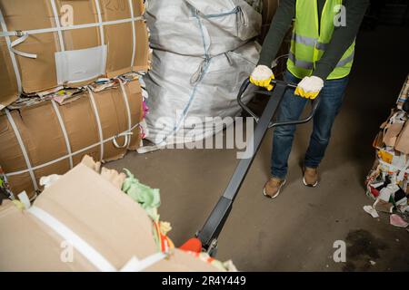 Vista ritagliata dello smistatore in gilet e guanti ad alta visibilità utilizzando transpallet manuale e spostando la carta di scarto nel centro di smistamento rifiuti, smistamento rifiuti e. Foto Stock