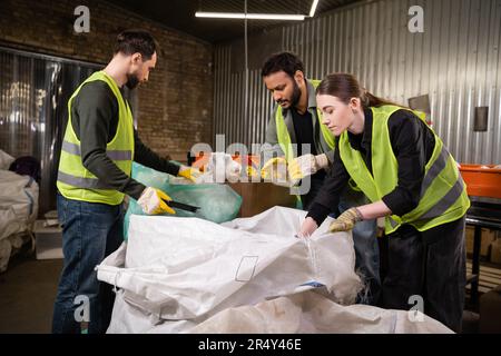Lavoratori multietnici in giubbotti di sicurezza e guanti di protezione che separano i rifiuti vicino ai sacchi mentre lavorano insieme in stazione di smaltimento rifiuti, spazzatura Foto Stock