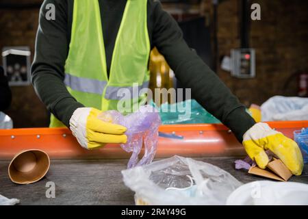 Vista ritagliata dello smistatore nei guanti protettivi e nel giubbotto che preleva i rifiuti dal trasportatore mentre si lavora in un centro di smistamento dei rifiuti sfocato, smistamento dei rifiuti e. Foto Stock