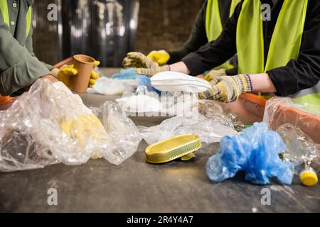 Vista ritagliata dei lavoratori in giubbotti e guanti ad alta visibilità che separano i diversi rifiuti di plastica e carta sul trasportatore mentre si lavora nello smaltimento dei rifiuti Foto Stock