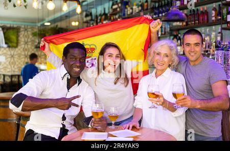 Felici tifosi multirazziali che tengono e si avvolgono in una bandiera di Spagna e sostengono la squadra nazionale di calcio nel bar Foto Stock