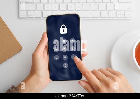 Protezione della privacy. Donna che immette il codice pin sullo smartphone al tavolo bianco, vista dall'alto. Blocco e tastierino numerico sullo schermo Foto Stock