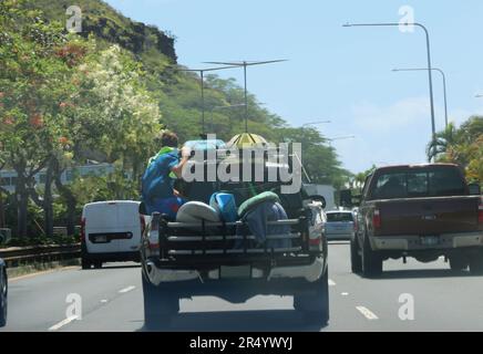 Honolulu, HI, USA - 30 maggio 2023. Il ragazzo cavalca in piedi sul retro aperto di un camion che tiene le tavole da surf su una strada trafficata. Foto Stock