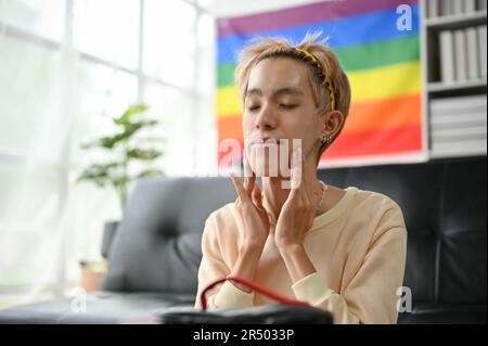 Un giovane uomo gay asiatico felice e calmo sta applicando il trattamento viso crema idratante o siero e massaggiando il viso prima di applicare il trucco. Foto Stock