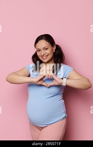 Donna incinta gioiosa gesta con le mani, mostrando la forma del cuore con le dita, sorride guardando la macchina fotografica, isolato sul rosa Foto Stock