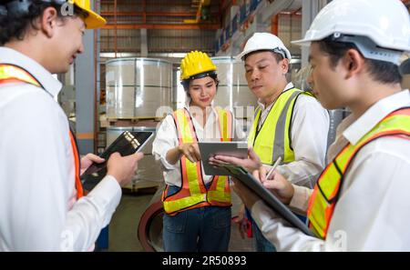 Il Senior Engineer Manager forma i nuovi dipendenti all'interno della fabbrica di lamiere. Tutti indossano giubbotto di sicurezza e elmetto. La pila di rulli in lamiera di acciaio si trova in Foto Stock