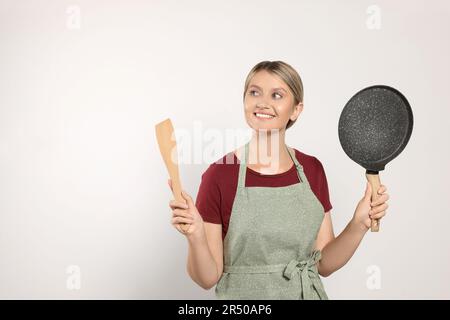 Bella giovane donna in grembiule pulito con utensili da cucina su sfondo grigio chiaro Foto Stock