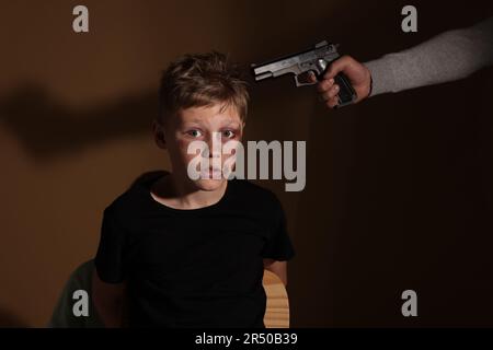 Kidnapper puntando la pistola al ragazzino preso in ostaggio su sfondo scuro Foto Stock