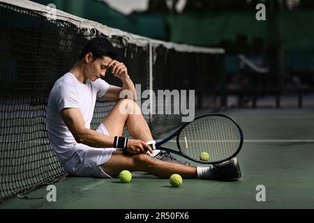 Vista laterale di un giocatore di tennis maschile asiatico deluso seduto sul campo, sconvolto da perdita di concorrenza, emozioni di fallimento Foto Stock