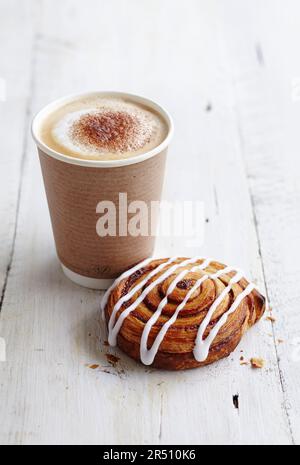 Cappuccino da asporto schiumoso accanto a una pasta frolla alla cannella cosparsa di glassa Foto Stock