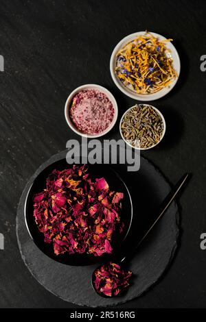 Fiori di rosa in ciotola nera, fiori di lavanda, sale floreale e mix di fiori in ciotole bianche su piatto di ardesia Foto Stock