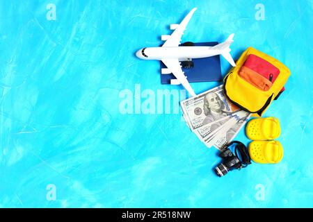 Piccola borsa piena di contanti, passaporto, modello di aeroplano, macchina fotografica giocattolo e scarpe da spiaggia poste su un accattivante sfondo blu con spazio copia. Viaggi essen Foto Stock