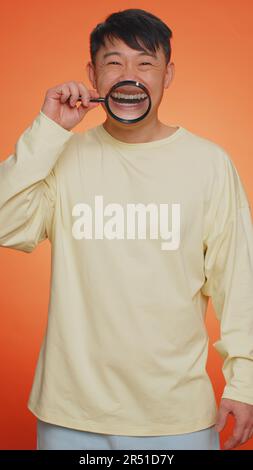 Uomo asiatico che tiene lente di ingrandimento su denti sani bianchi, guardando la fotocamera con espressione felice, mostrando divertente sciocco viso sorridente bocca. Ragazzo cinese isolato su sfondo arancione. Verticale Foto Stock
