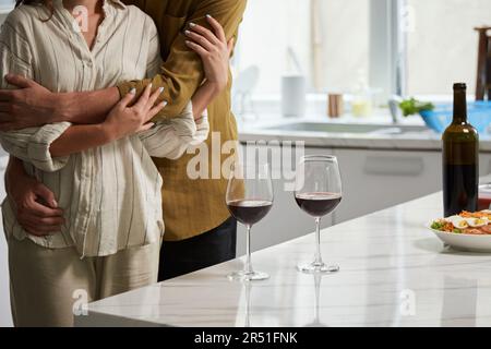 Immagine ritagliata di una coppia agghiacciante in piedi al bancone della cucina con bicchieri da vino Foto Stock