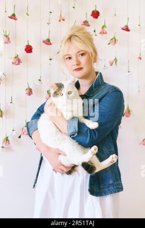 Studio ritratto di bella giovane adolescente 15 - ragazza di 16 anni che indossa giacca denim, posa su sfondo bianco con fiori appesi, tenendo carino gatto, Foto Stock