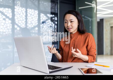 Felice donna d'affari asiatica che lavora in un ufficio moderno utilizzando un computer portatile per videochiamare e riunioni online con colleghi di lavoro, donna sorridente e divertente dando una presentazione. Foto Stock