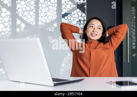 Felice e sorridente donna asiatica di successo ha finito il lavoro soddisfatto con il risultato del lavoro e la realizzazione, le mani dietro la testa e sorridente primo piano Foto Stock