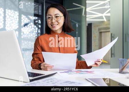 Giovane finanziatore asiatico di successo dietro il lavoro di carta all'interno dell'ufficio, commercialista donna d'affari che esamina i documenti contrattuali e le relazioni sorridente e felice, soddisfatto con i risultati di realizzazione utilizzando il portatile. Foto Stock