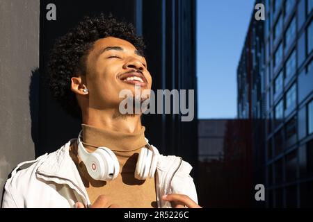 Giovane afroamericano allegro con acconciatura afro e occhi chiusi appoggiati a una parete con cuffie intorno al collo Foto Stock