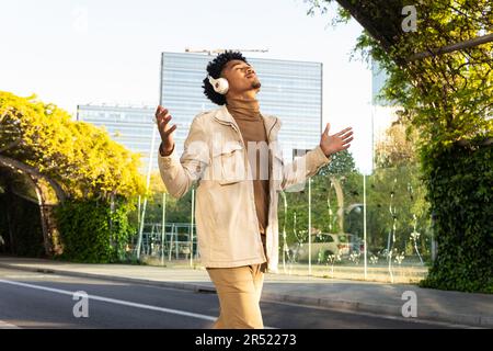 Felice afro americano giovane con gli occhi chiusi ascoltando musica con cuffie e braccia aperte mentre cammina per la strada contro gli alberi Foto Stock