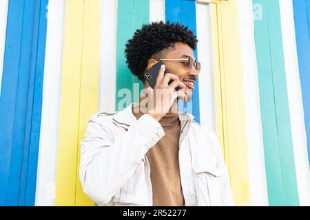 Positivo afro americano giovane con occhiali da sole pettinato afro parlando sul suo cellulare e sorridendo mentre guardando lontano contro un muro Foto Stock