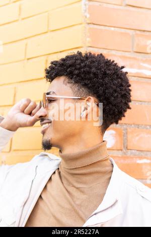Giovane aragosta afroamericana in dolcevita beige e occhiali da sole con capelli ricci che si guardano via mentre si appoggiano contro un edificio in mattoni sulla strada Foto Stock