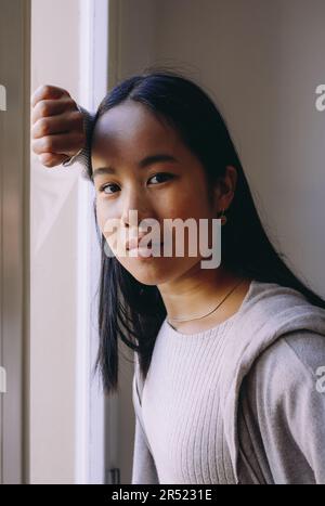 Ritratto di seria giovane donna asiatica in abiti casual in piedi vicino alla finestra e guardando la fotocamera in luce del giorno Foto Stock