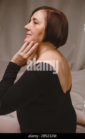 Vista laterale della ballerina femminile matura in leotard nero con i capelli corti marroni che toccano la guancia e che guardano via contro il tessuto beige in studio Foto Stock