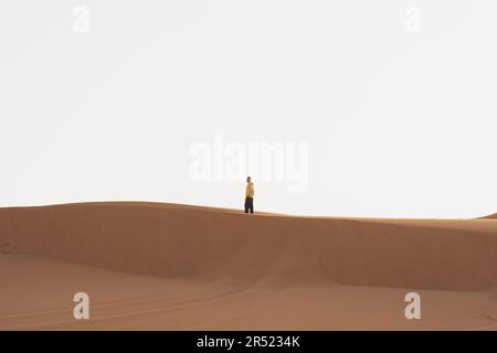 Vista laterale di una persona distante che cammina su dune sabbiose contro il cielo nuvoloso della città di Marrakech durante il viaggio estivo attraverso il Marocco Foto Stock