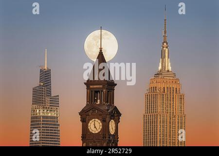 La luna piena sorge dietro la Torre dell'Orologio di Lackawanna a Hoboken, New Jersey, accanto all'iconico Empire state Building e al 1 Vanderbilt Foto Stock